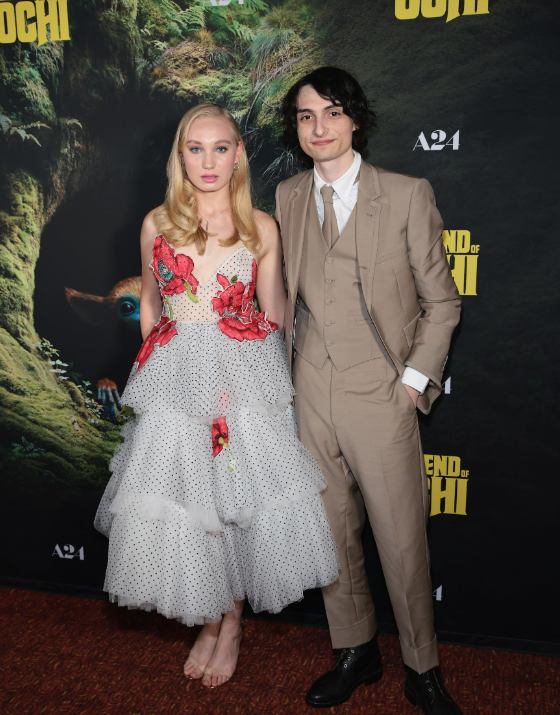 Duas pessoas posando para foto em um evento, com um fundo colorido e a imagem de um personagem de um filme.